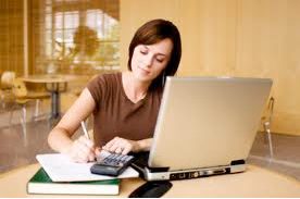 A woman sits at a desk using a laptop and calculator while writing notes in a notebook to improve her study habits in a well-lit room.