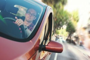 A man driving a red car, viewed through the side mirror, focused on the road ahead with blurred street surroundings.