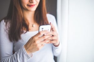 A woman with brown hair, smiling slightly, looking at her smartphone, wearing a white shirt and standing by a window.