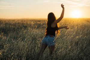 Woman with long hair, wearing shorts and a tank top, raising her arm in a wheat field at sunset.