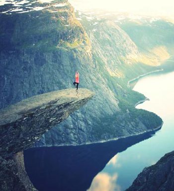 A person stands on the edge of a steep cliff overlooking a misty fjord at sunrise.