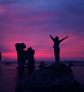 A silhouette of a person with arms raised stands on a rock against a vivid pink and purple sunset sky over the sea.
