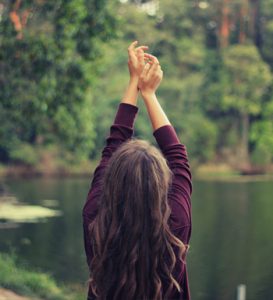 A woman with long hair stretches her arms upward by a serene lake, surrounded by trees.