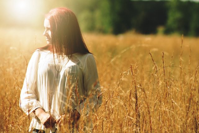 A woman with red hair standing in a golden field, looking to the side as sunlight streams through her hair.