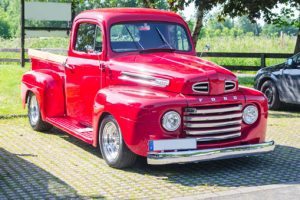 Red vintage ford pickup truck parked on a cobblestone area with green trees in the background.