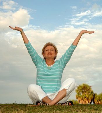 A woman in a striped shirt and white pants sits cross-legged on grass, arms raised joyfully, with a cloudy sky in the background.