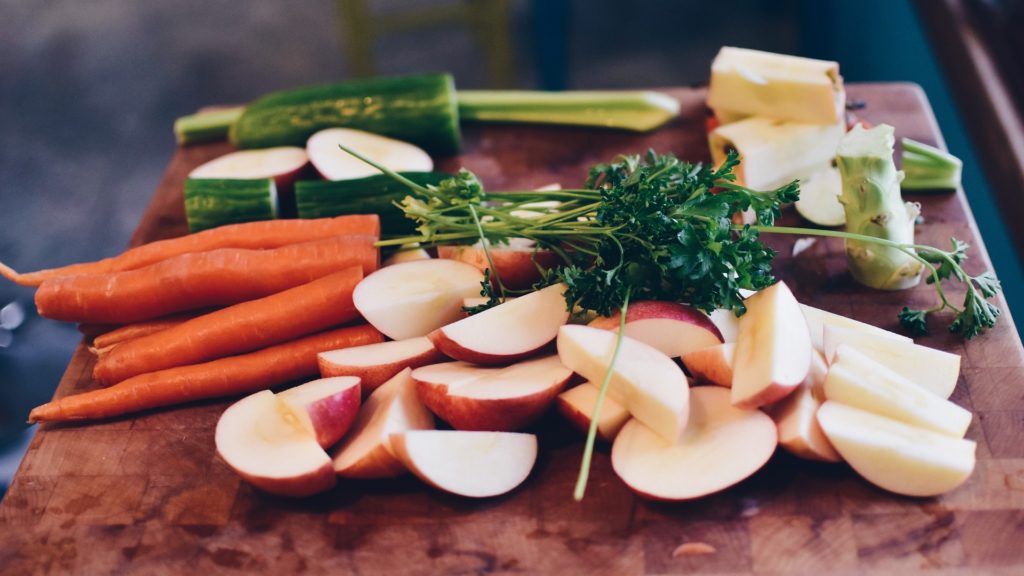 A variety of freshly chopped vegetables and fruits, including carrots, cucumbers, apples, and parsley, on a wooden cutting board.