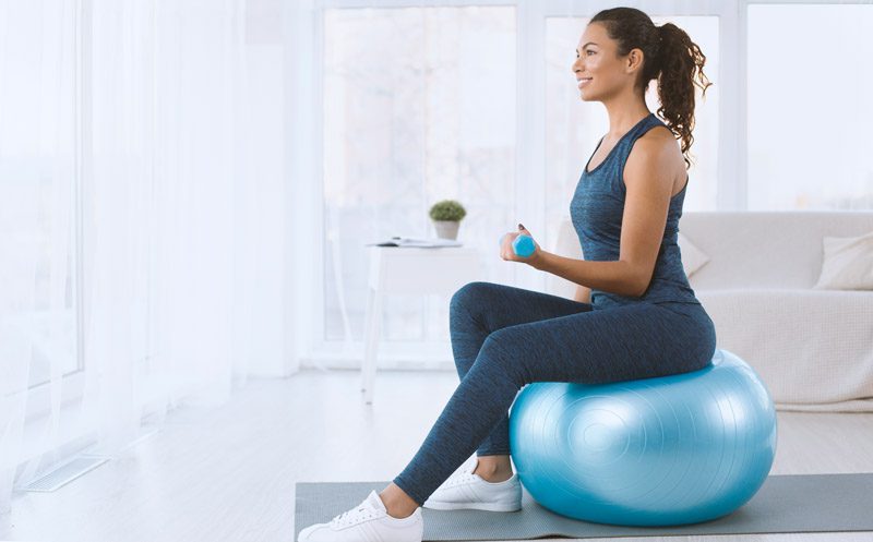 A woman in blue activewear sits on a blue exercise ball holding a small weight, smiling in a bright, modern living room.