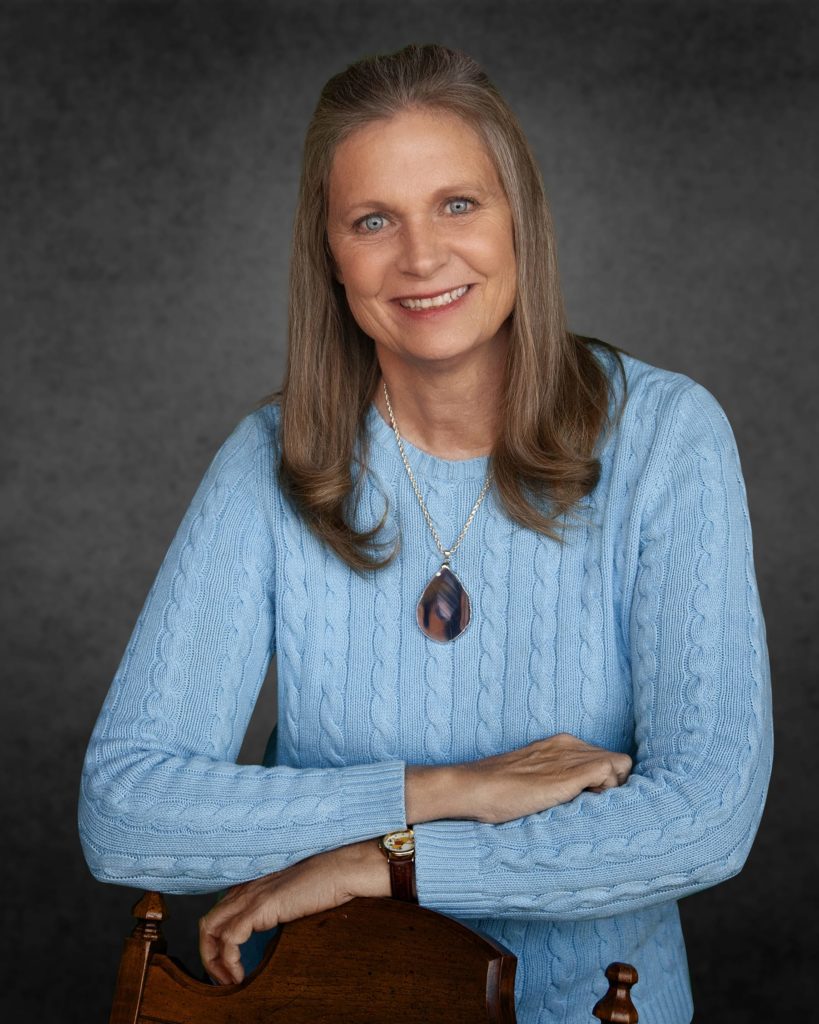 Portrait of a smiling middle-aged woman in a blue sweater, leaning on a wooden chair, wearing a pendant necklace.