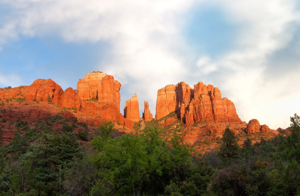 Red rock formations illuminated by the setting sun in sedona, with green foliage in the foreground and a clear blue sky overhead.