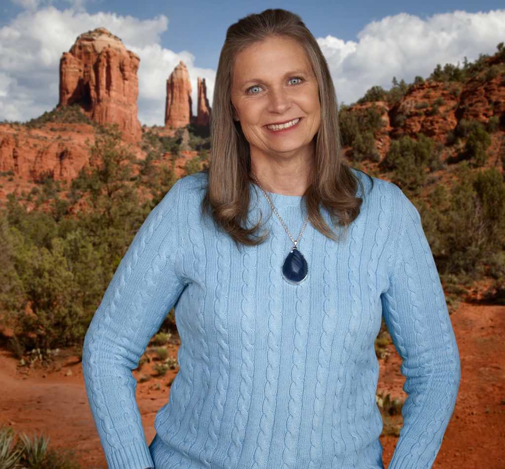 Woman in a blue sweater smiling, standing in front of a scenic desert landscape with red rock formations.