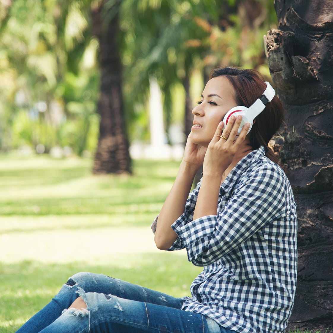 A woman in a plaid shirt and jeans sits by a tree, enjoying music on her headphones, in a sunny park.