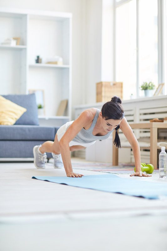 A woman in athletic wear doing a push-up on a yoga mat in a bright living room, with a water bottle beside her.