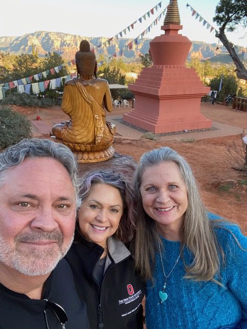 Three people smiling for a selfie with a large buddha statue and a stupa in a serene park setting with trees and hypnotherapy techniques for weight loss flags.