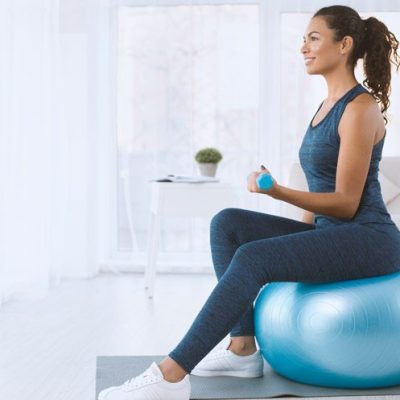 A woman in blue activewear sits on a blue exercise ball holding a small weight, smiling in a bright, modern living room.