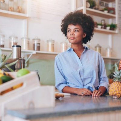 A woman with curly hair leans on a kitchen counter, looking thoughtful, surrounded by fresh fruits in baskets.