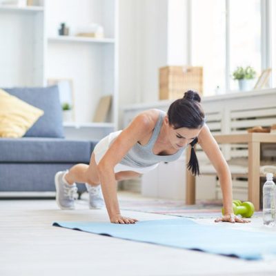 A woman in workout gear performs a push-up on a yoga mat in a brightly lit living room with shelves and a sofa in the background.