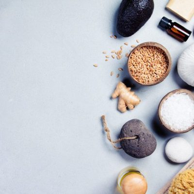 Flat lay of assorted spa products including avocados, sea salt, stones, and essential oils on a light background.