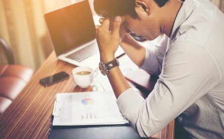 A stressed businessman holding his head at a desk with a laptop, papers with charts, and a coffee cup nearby.